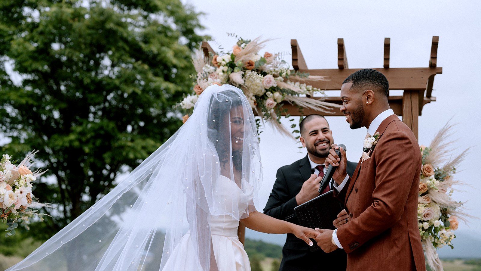 A bride in a pearl veil and a groom in a rust suit hold hands and laugh while Matt Perry speaks into a microphone beneath a floral arch